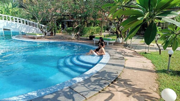 A swimming pool with a white bridge, surrounded by greenery and a paved walkway. - Sashas Holiday Village (Vagator)