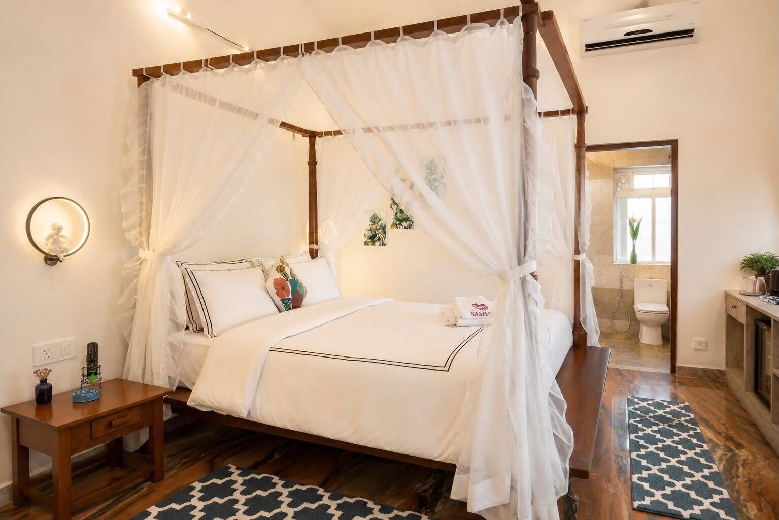 A bedroom with a four-poster bed, a bedside table, a nightstand, a wall-mounted lamp, and a bathroom visible through an open door.