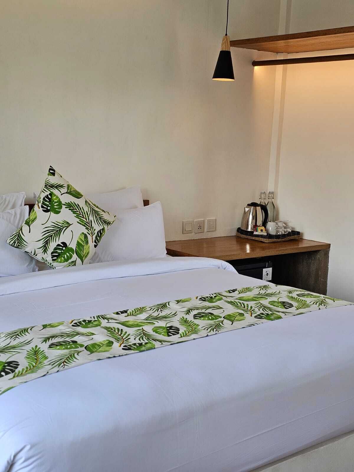 Een opgemaakt bed met witte lakens en een sprei met een bladmotief. Er staat een houten nachtkastje met een waterkoker en glazen naast het bed.