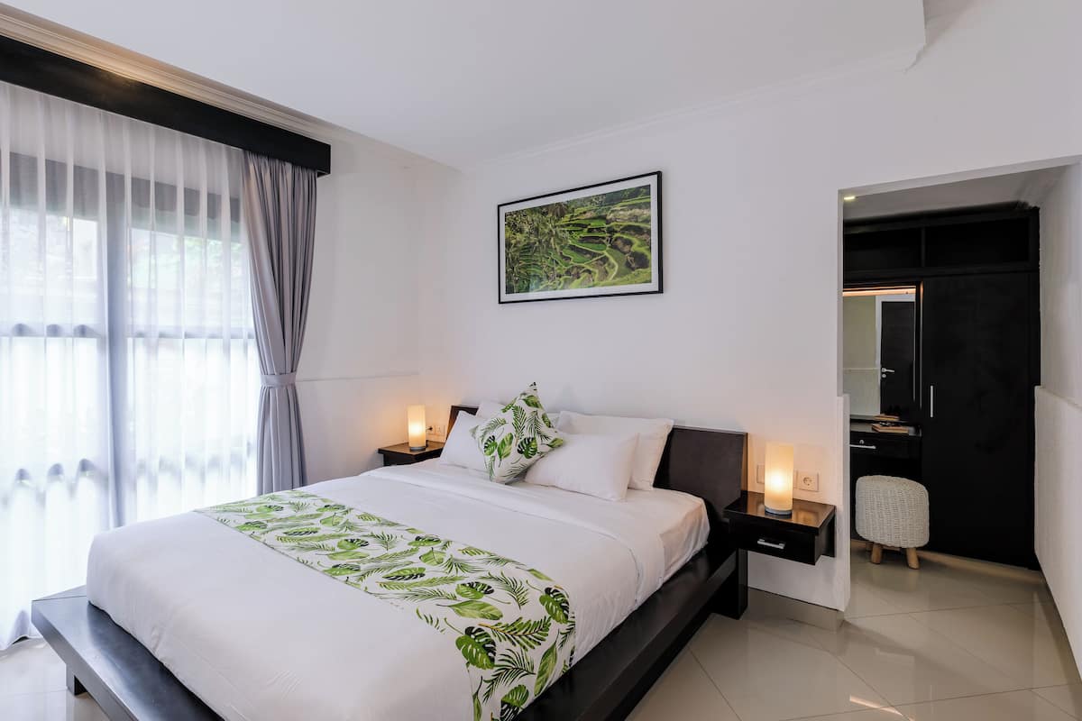 A modern bedroom with a large bed, a nightstand with a lamp, a framed picture on the wall, and a view into a bathroom.