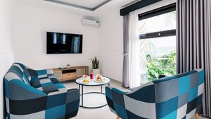 A modern living room with two sofas, a TV, a small table with food, and a large window with curtains. - Chandi Hotel Ubud  (Ubud)