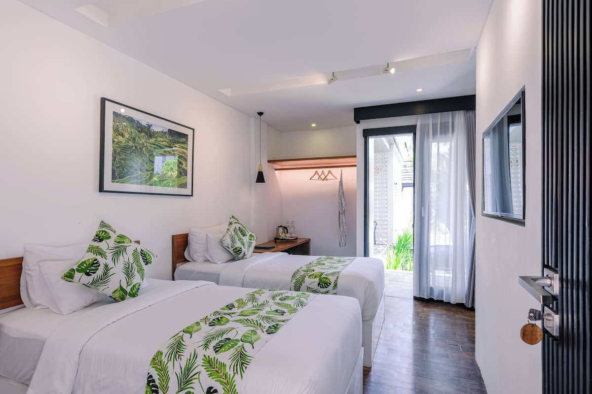A hotel room with two beds, a large window with curtains, a framed picture on the wall, and a small table with a lamp.