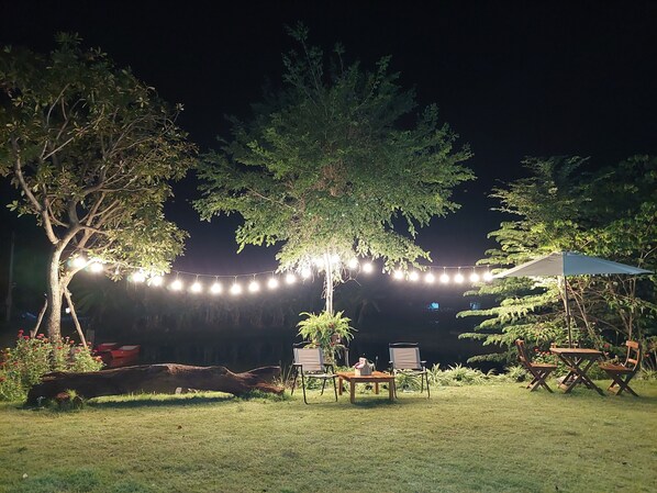 Un patio al aire libre con luces colgantes, una mesa con sillas y un árbol grande que da sombra.
