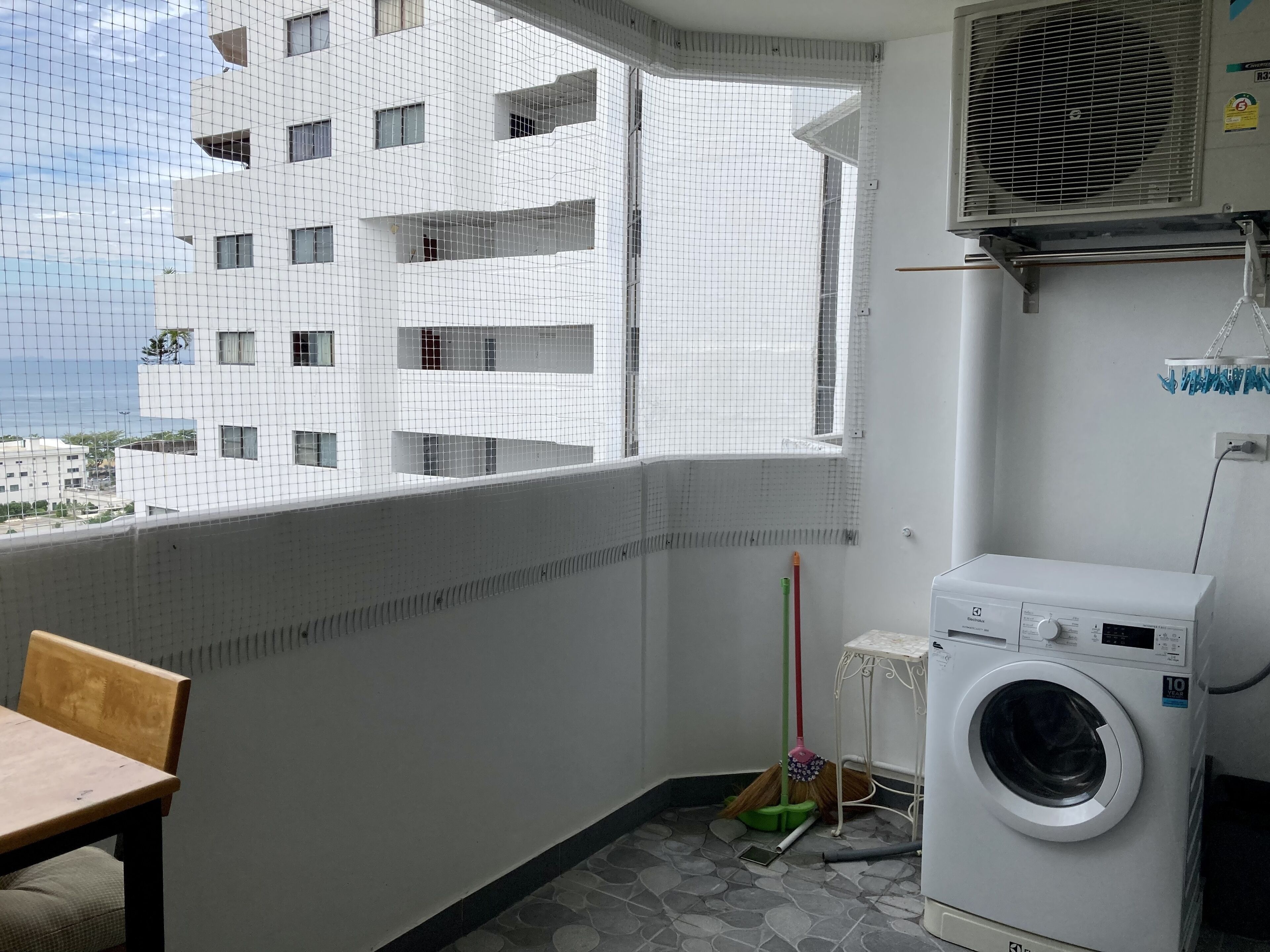 A balcony with a view of a white building, a washing machine, and a broom.