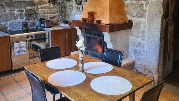 A rustic kitchen with a stone wall, a wood-burning stove, and a dining area with a wooden table and chairs. - ¡A 10 Minutos de Santiago de Compostela! Casa Ideal Para Familias (la esclavitud,Padron)