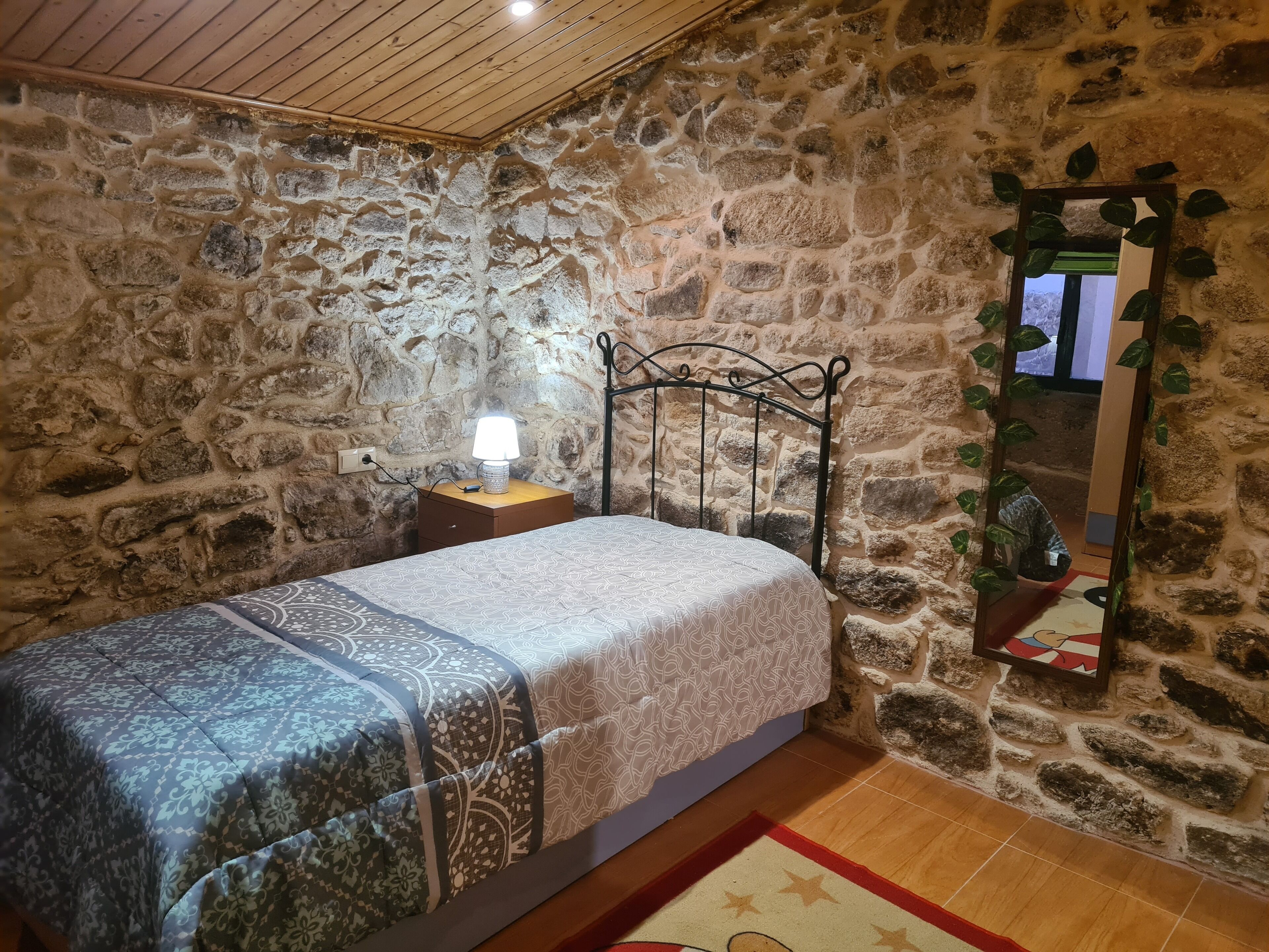 Een slaapkamer met een stenen muur, houten plafond, een bed met behang, nachtkastje met lamp en spiegel met planten.
