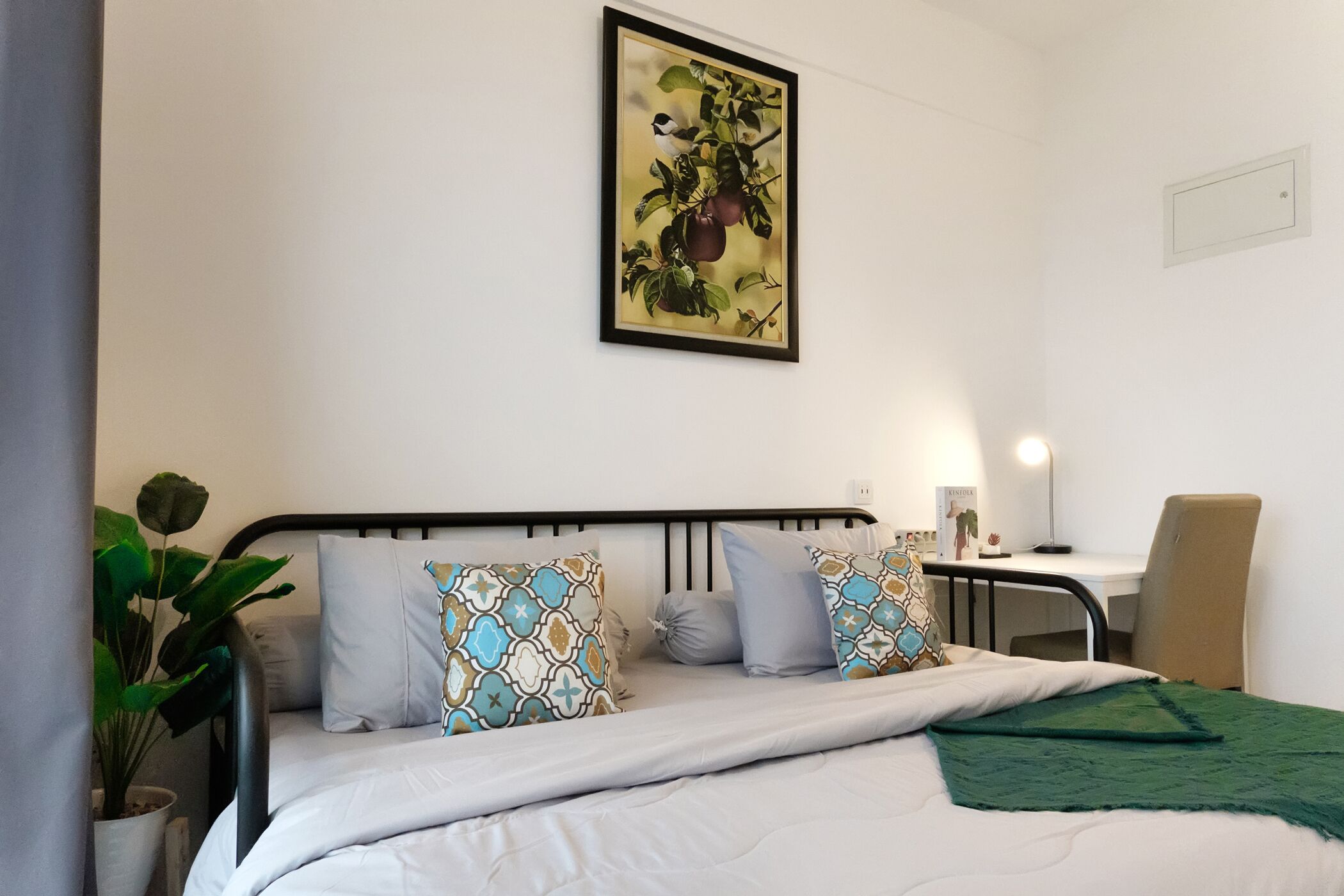 A neatly made bed with patterned pillows, a green throw blanket, and a wall-mounted picture of fruit.