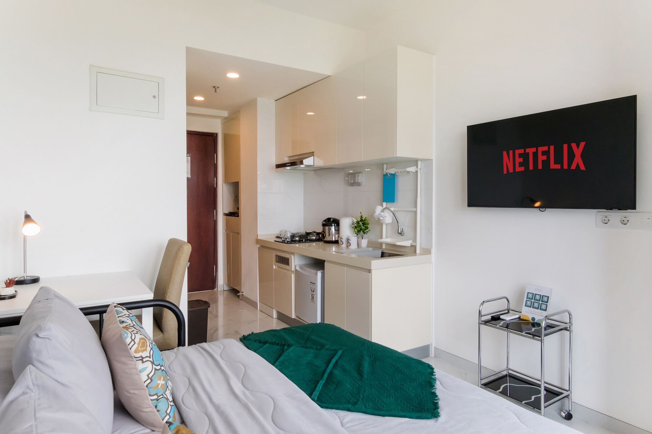 A modern hotel room with a bed, a desk, a chair, a kitchenette, and a TV displaying the Netflix logo.