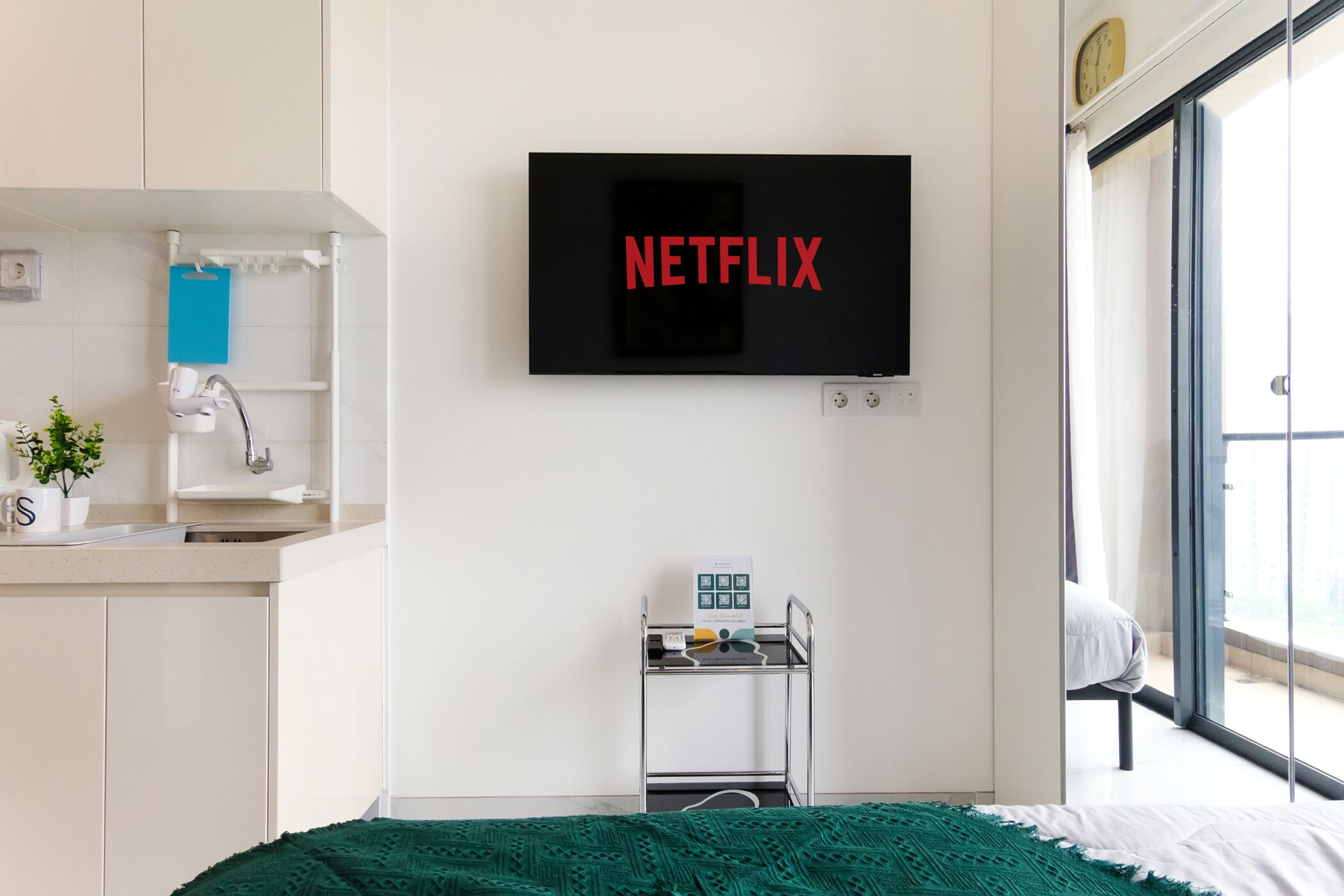 Hotelkamer met een tv waarop 'NETFLIX' staat, een klein tafeltje met een klok, een raam met uitzicht en een bed met een groene deken.