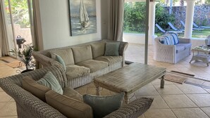 Un vaste espace de vie agrémenté de meubles en osier, d’un tableau représentant un voilier et offrant une vue sur un jardin et une piscine.