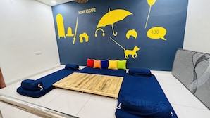 A room with a blue bed, colorful pillows, a wooden platform, and a wall mural with silhouettes of a hat, umbrella, headphones, a dog, and a speech bubble with the number 100. - Home Escape Luxe (Ujjain)