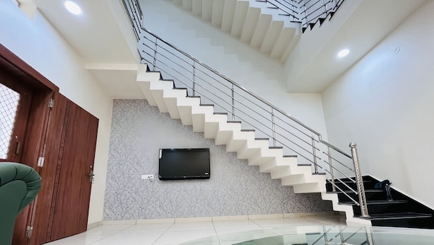 A modern interior with a staircase, a flat-screen TV mounted on a marble wall, and a glass railing in the foreground. - Home Escape Luxe (Ujjain)