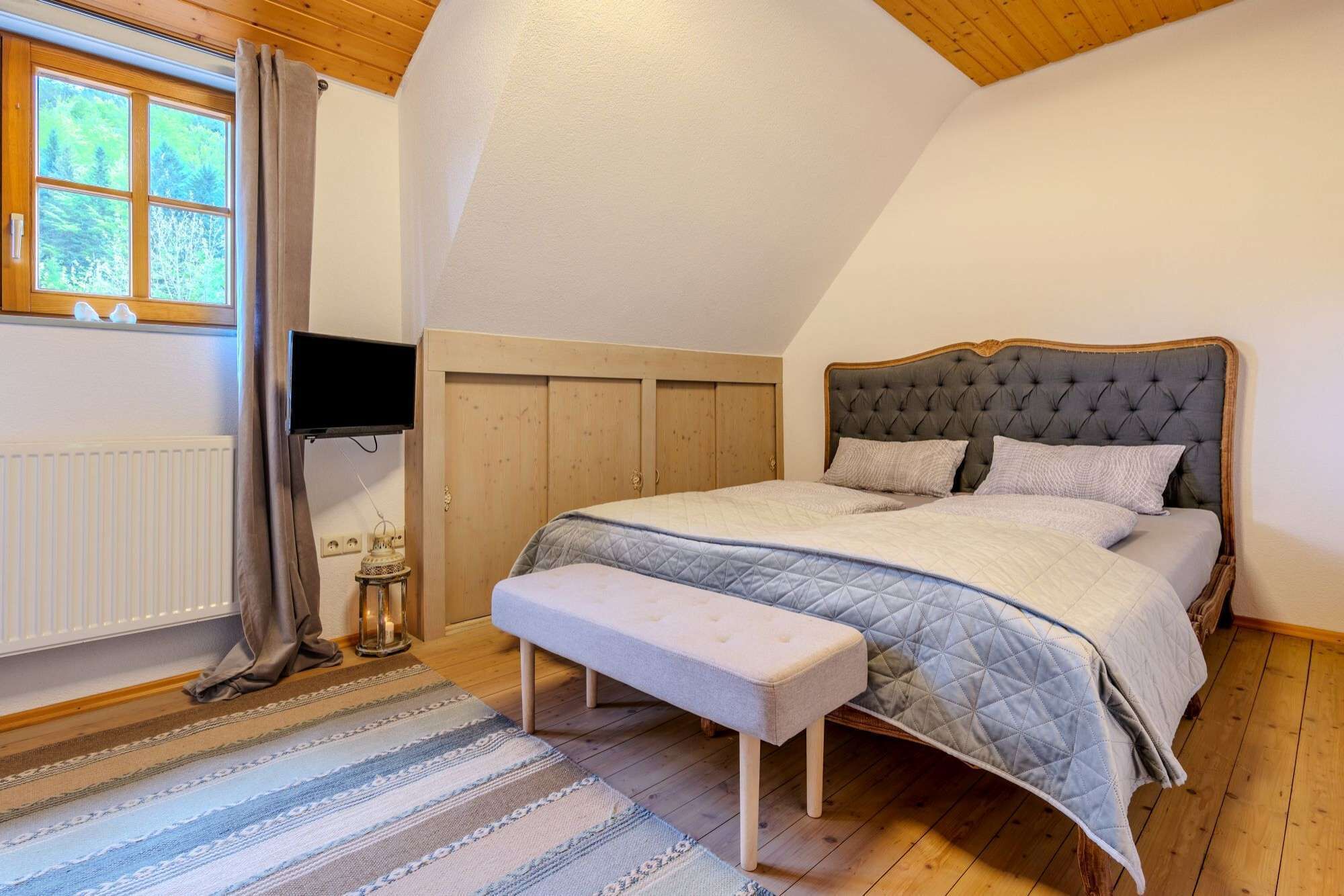 A bedroom with a large bed, a bench, a TV, and a window with curtains.