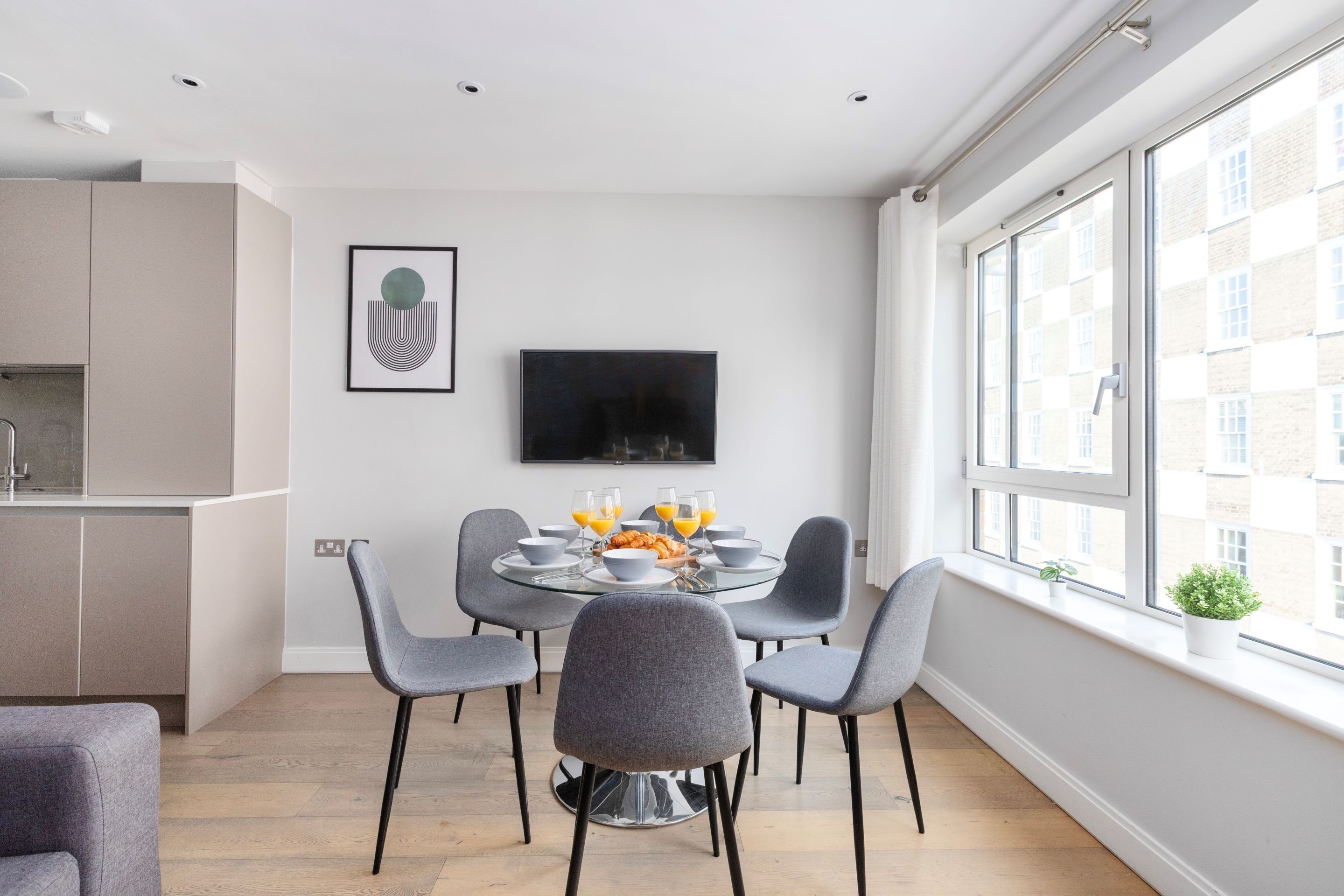 A modern dining area with a round glass table, grey upholstered chairs, and a flat-screen TV mounted on the wall.