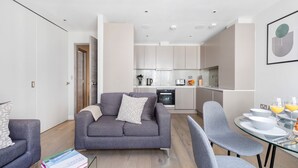 Ein modernes Apartment mit einem grauen Sofa, einem Glastisch und einer Küche im Hintergrund.
