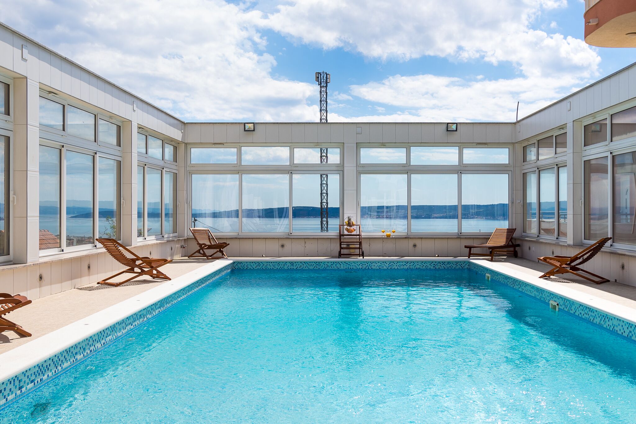 An indoor swimming pool with clear views of the sea and mountains, surrounded by large windows and sun loungers.