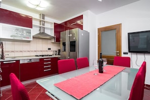 A modern kitchen with red cabinets, stainless steel appliances, a glass dining table with a pink placemat, and red chairs.