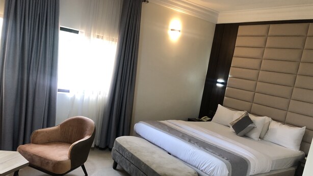 A hotel room with a bed, a chair, a bedside table, and a nightstand with a phone. - Roban Hotels Limited (Enugu)