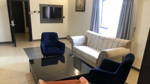 A modern living room with a black glass coffee table, two blue armchairs, a white sofa, and a small orange ottoman. - Roban Hotels Limited (Enugu)