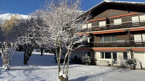 A two-story building with a balcony, surrounded by snow-covered trees and a mountain in the background. - Ferienwohnung Waldrand EG by Interhome (Lenk)