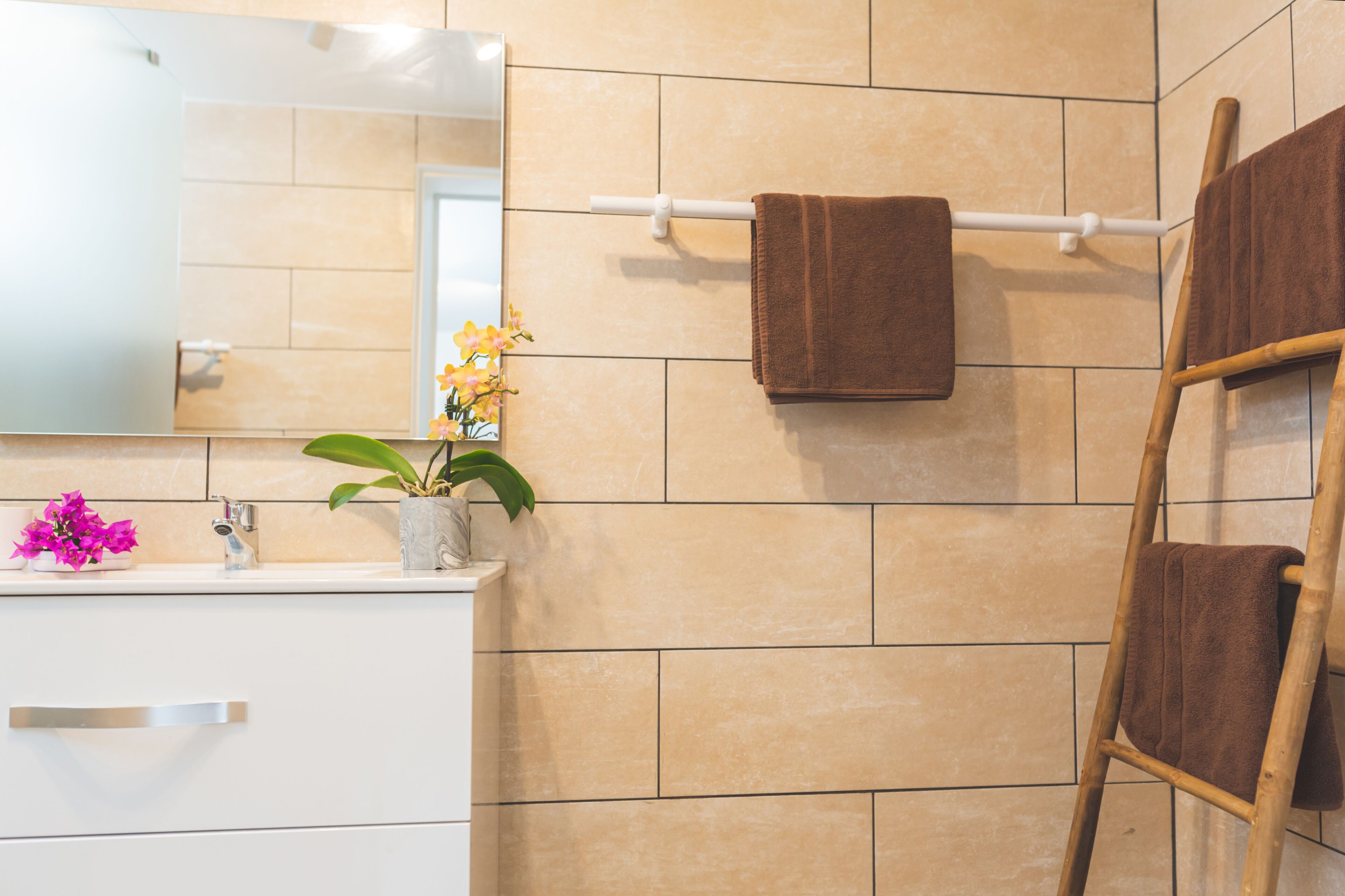 Een badkamer met beige tegels, een witte wastafel, een spiegel, een handdoekhouder met twee handdoeken, een plant in pot en een houten ladder.
