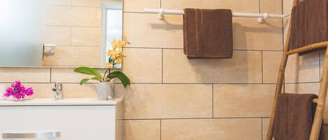 Een badkamer met beige tegels, een witte wastafel, een spiegel, een handdoekhouder met twee handdoeken, een plant in pot en een houten ladder.