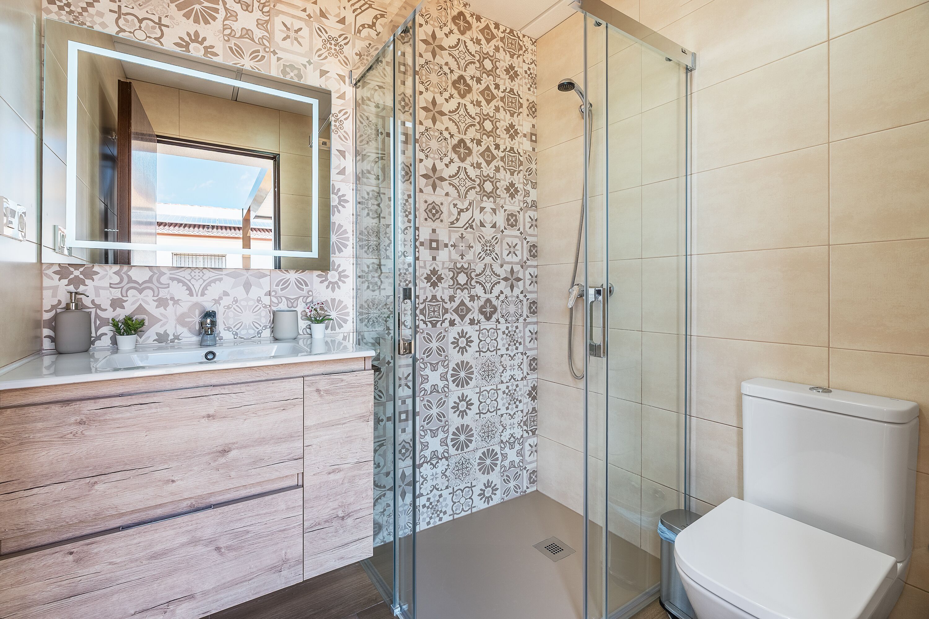 A bathroom with a glass shower enclosure, a toilet, a sink with a mirror, and patterned tiles.
