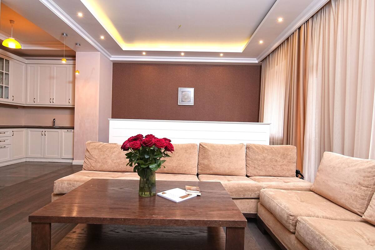 Una sala de estar moderna con un sofá, una mesa de centro con un jarrón de rosas rojas, y una cocina al fondo.
