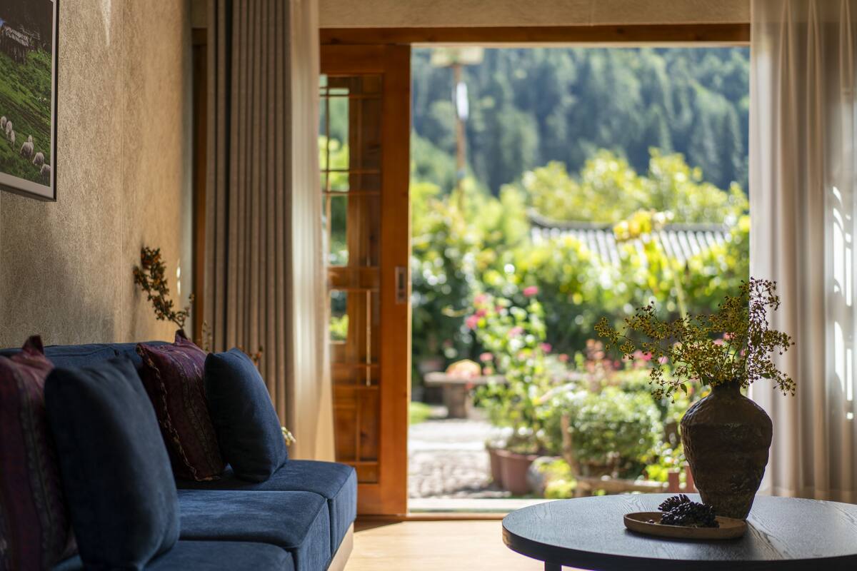 A room with a blue sofa, a wooden table with a vase and pine cones, and a view of a garden with potted plants and a wooden fence.