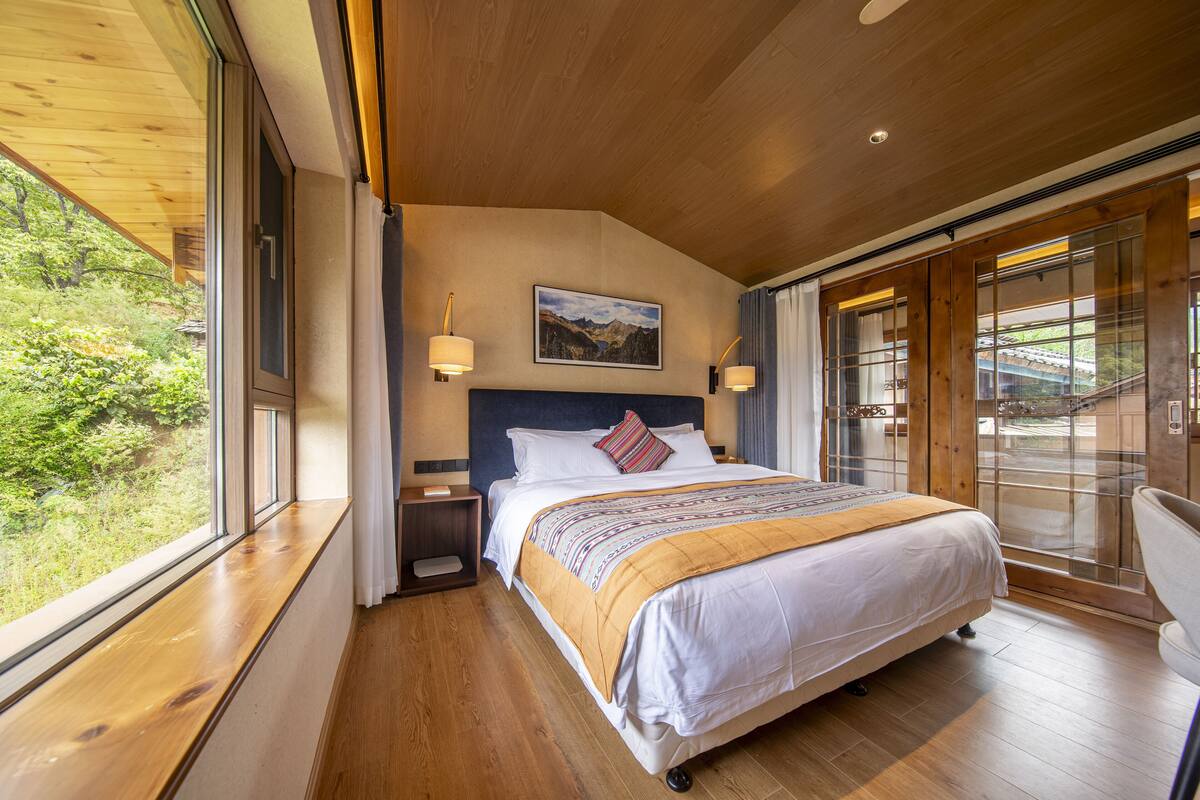 A bedroom with a large bed, wooden flooring, a nightstand, a wall-mounted lamp, and a window with a view of greenery.