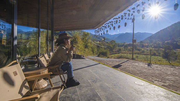 A person sitting on a bench under a wooden structure with a view of mountains and hanging decorations. - Shujiawuxiangluoguqingtubuminsu (Nujiang Lisu)