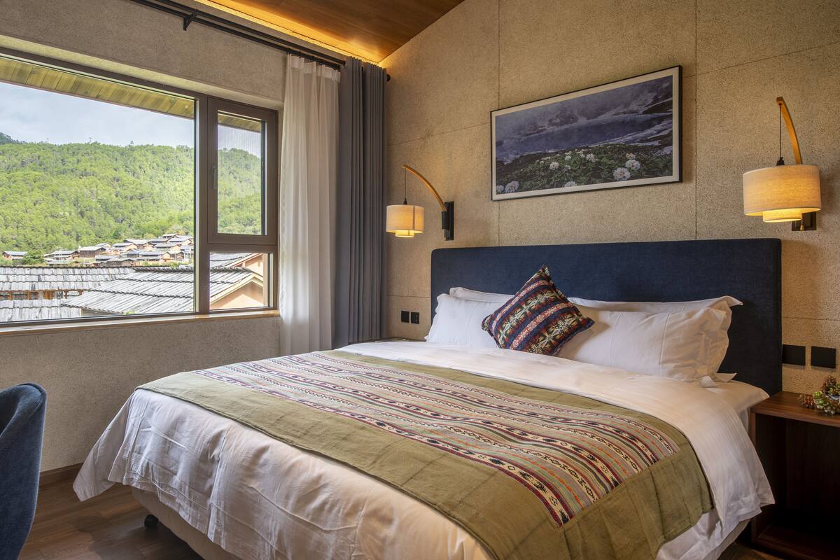 A hotel room with a large bed, a view of a mountainous landscape, a painting on the wall, and two wall-mounted lamps.