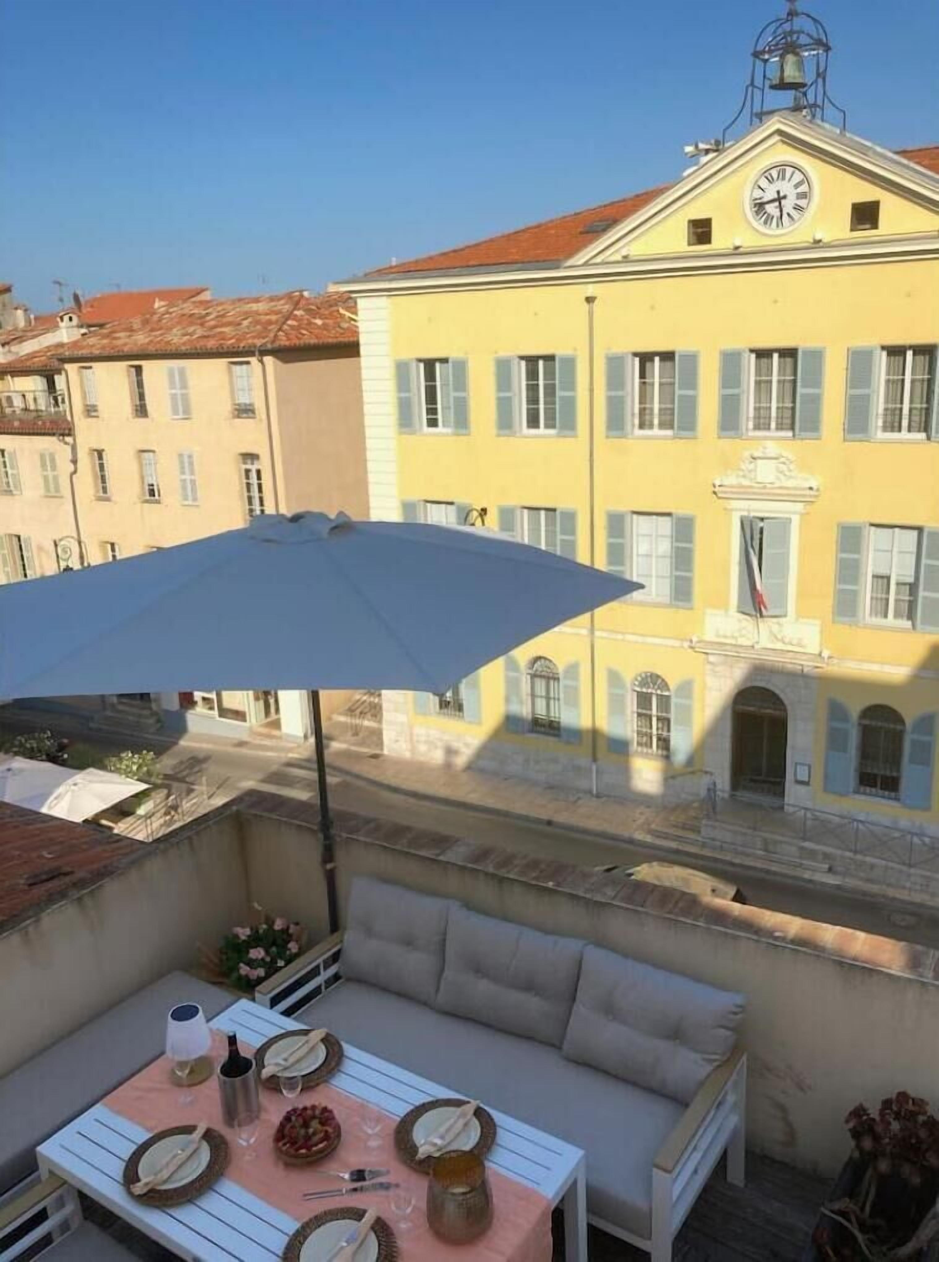 Un balcon avec une table dressée pour deux personnes, un canapé et une vue sur un immeuble jaune orné d’une horloge.