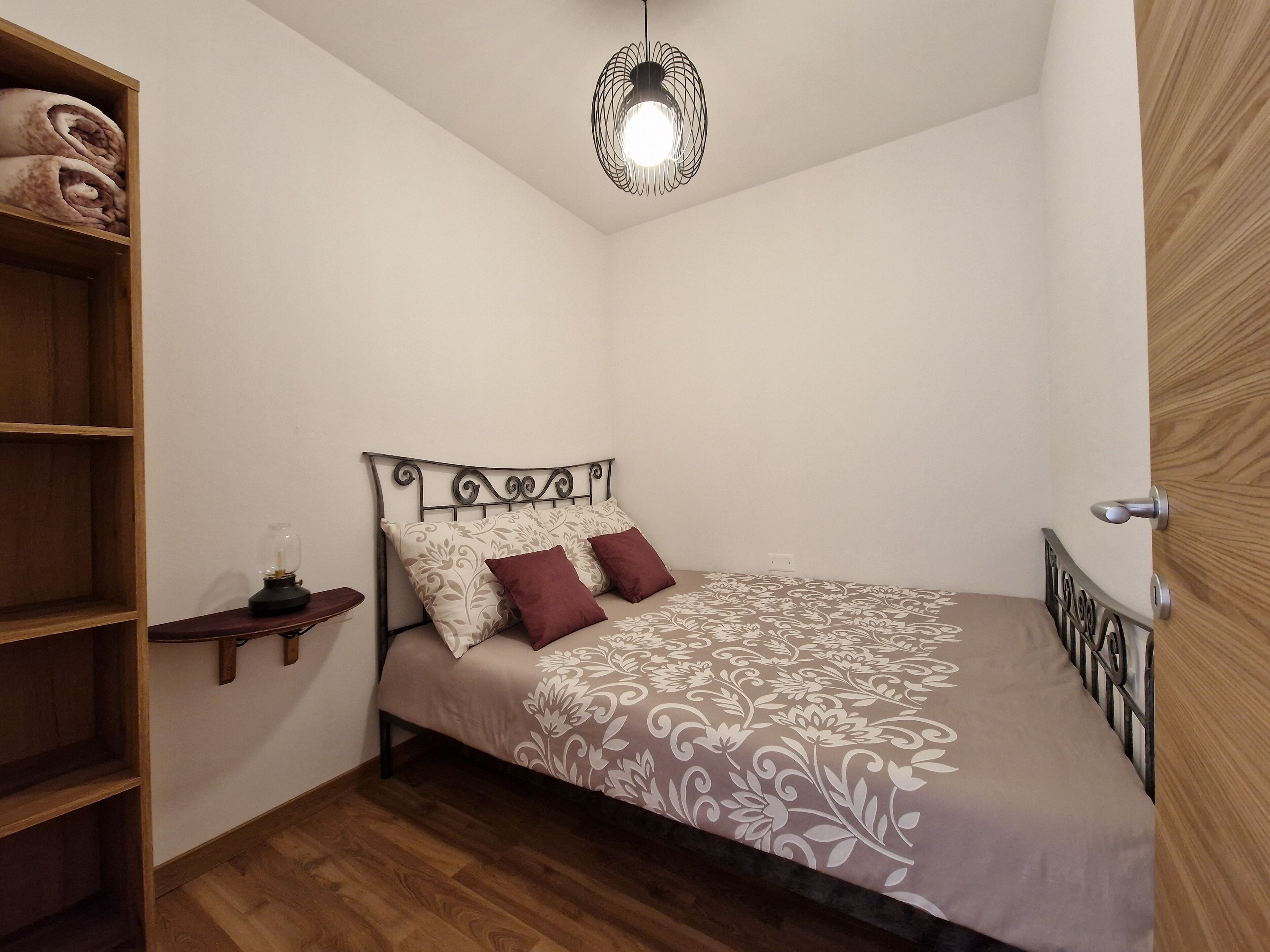A bedroom with a bed, a wooden shelf, a bedside table, and a hanging light fixture.