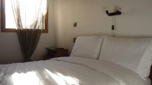 A bedroom with a bed, a bedside table, a wall-mounted light fixture, and a window with sheer curtains. - Nakou's House-Το σπίτι του Νάκου (Liopraso)