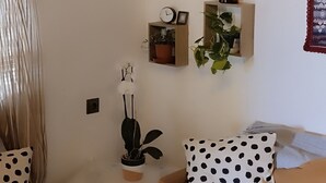 Un salon chaleureux avec un canapé, une table basse et une petite table d’appoint. On y trouve plusieurs plantes en pot, disposées sur des étagères, ainsi qu’une horloge murale.