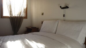 Une chambre à coucher avec un lit, une table de chevet, une lampe fixée au mur et une fenêtre laissant entrer la lumière du soleil.