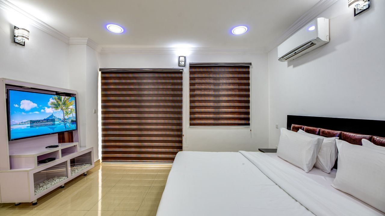 A hotel room with a bed, a TV, a shelf with items, and two windows with blinds.