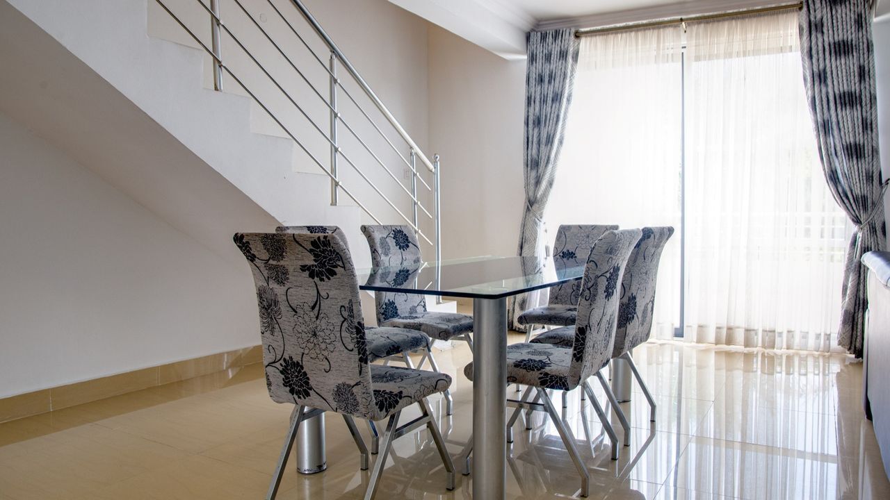 A dining area with a glass table and chairs, a staircase, and a view of the outside through sheer curtains.
