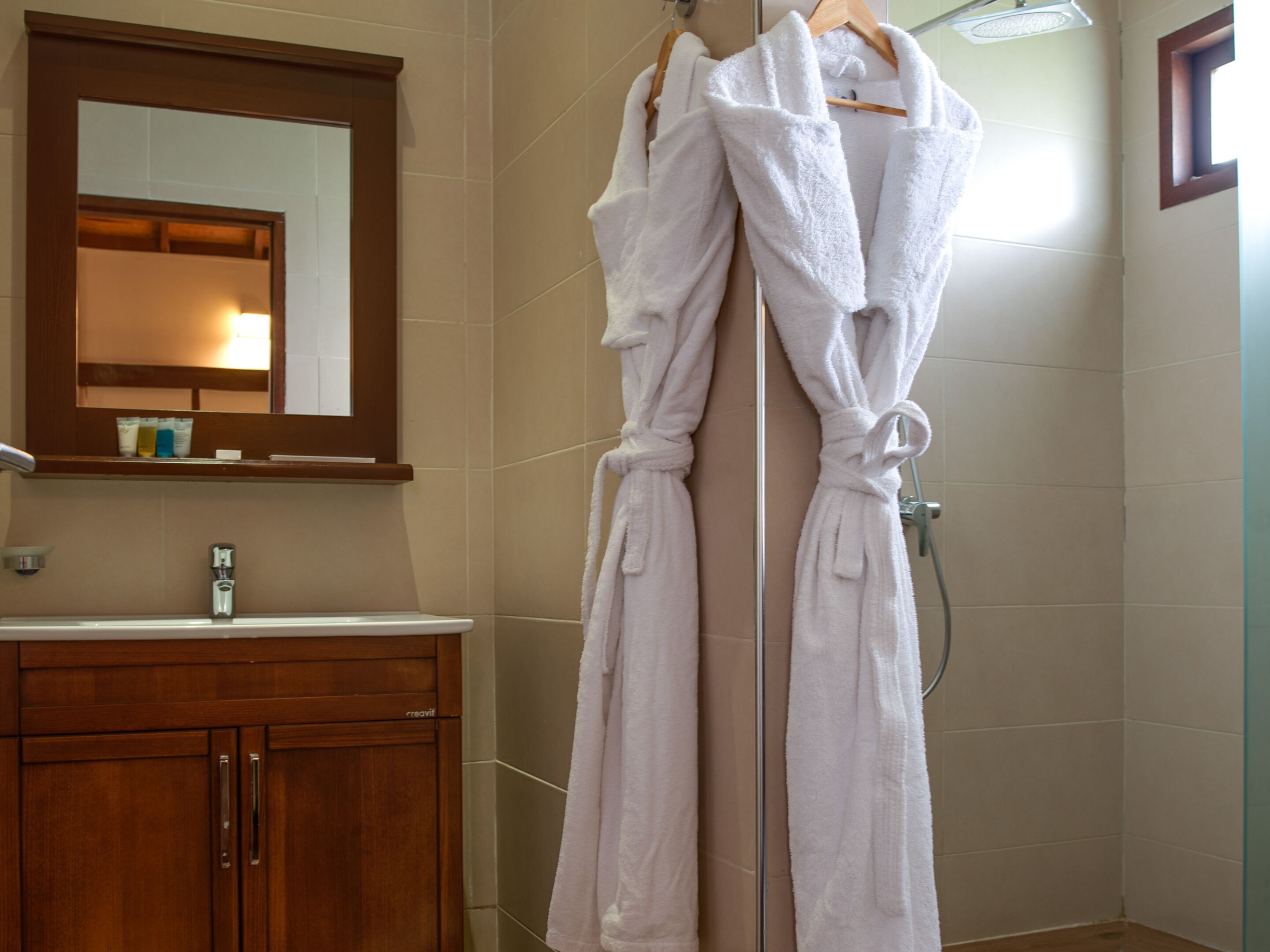 A bathroom with a wooden vanity, a mirror, and two white bathrobes hanging on a rack.