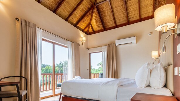 A bedroom with a bed, a chair, a wooden desk, a balcony with a railing, and a ceiling with a thatched roof. - Lakowe Lakes Hotel Golf and Country Estate by NEWMARK (Lekki)