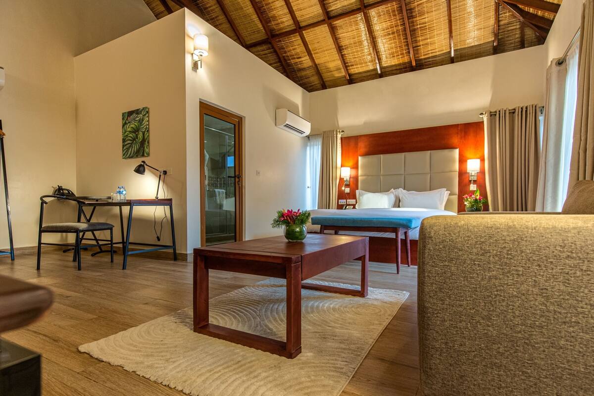Habitación de hotel con cama, sofá, escritorio y una silla. Se ve una ventana con cortinas, una puerta y un equipo de aire acondicionado en la pared.