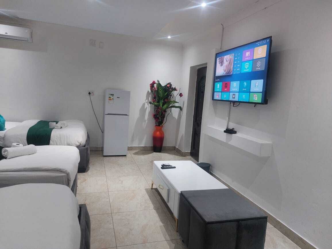 A hotel room with two beds, a small table, a TV mounted on the wall, a refrigerator, and a potted plant.