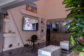 Un moderno vestíbulo de hotel con un televisor de pantalla plana montado en la pared, una recepción y un área de espera con mostrador y sillas.