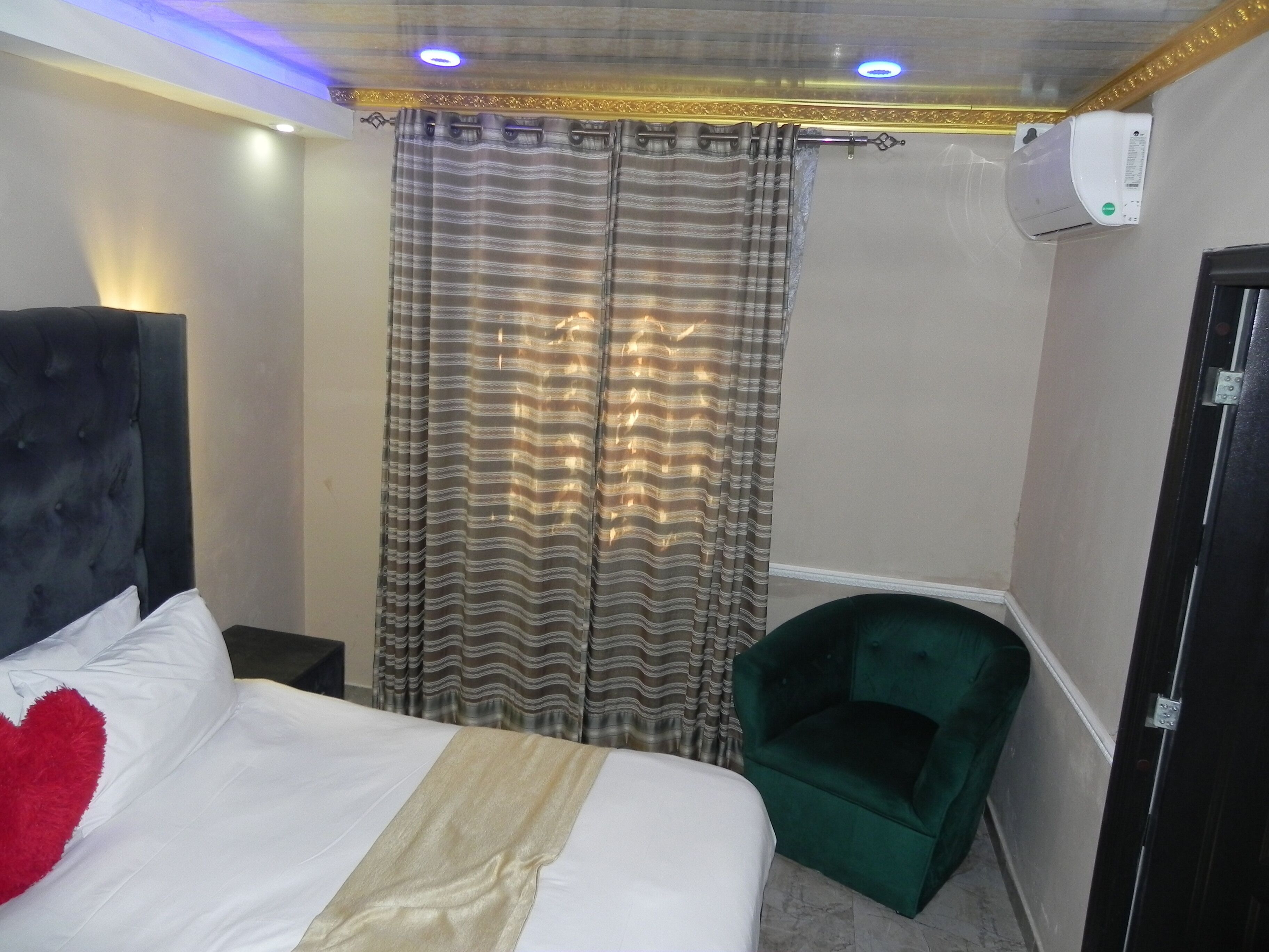 A hotel room with a bed, a green chair, a curtain, and an air conditioning unit.