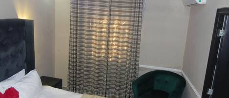 A hotel room with a bed, a green chair, a curtain, and an air conditioning unit.