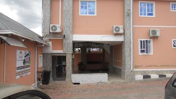 Entrada de un hotel con un área de espera techada, unidades de aire acondicionado y un cartel que indica tarifa para habitación individual.