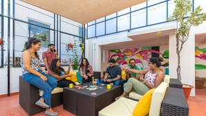 A group of people sitting on outdoor wicker furniture in a covered patio area with a view of a building through large windows. - Zostel Bangalore (Koramangala) (Bengaluru)