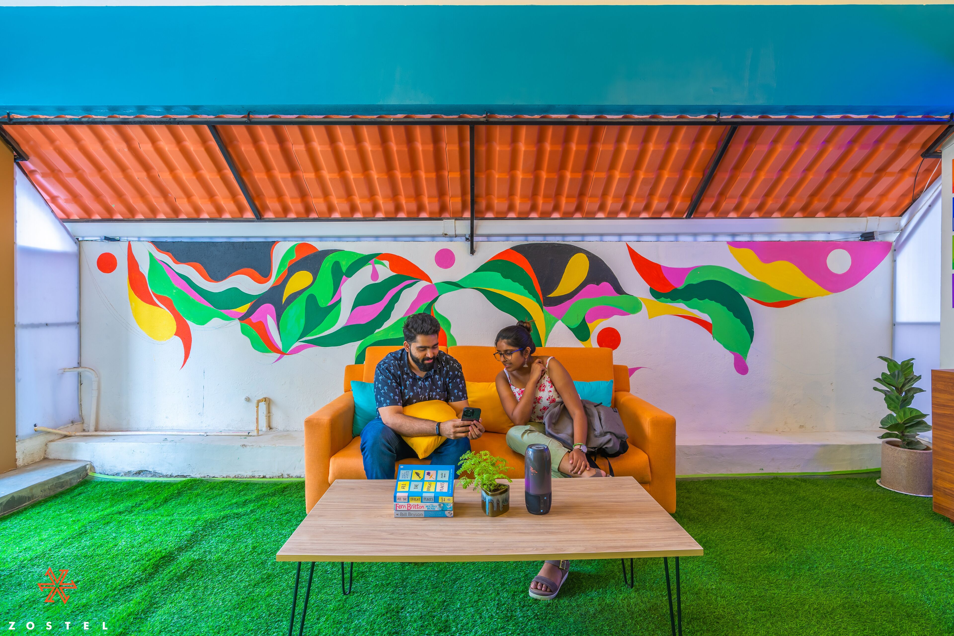 Quarto com um mural colorido, uma mesa de madeira com uma planta e livros, e um sofá com duas pessoas.