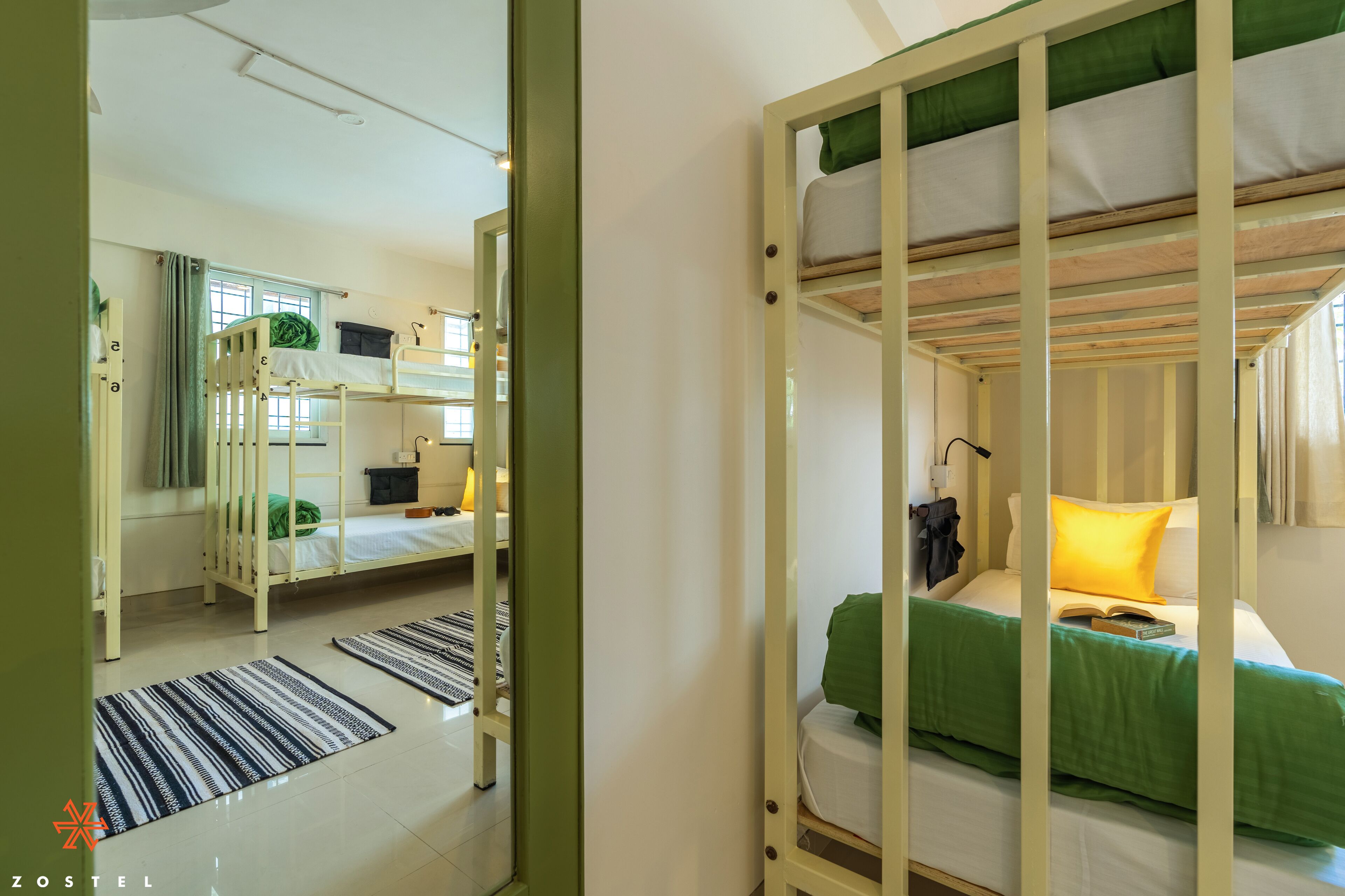 A dormitory room with bunk beds, a mirror, a window with curtains, and a green bedspread.